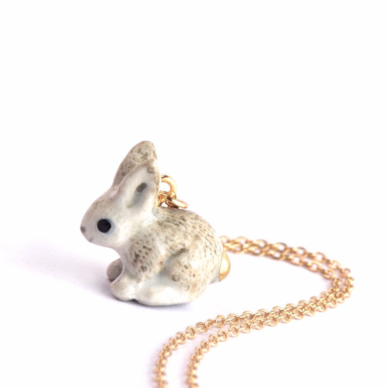 The Goldentail Rabbit Necklace showcases a small, delicate hand-painted porcelain bunny pendant with a speckled gray and white design. The bunny, in a curled position, features a golden tail. The heirloom quality piece hangs on a partially coiled golden chain against a plain white background.