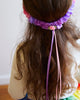 A young child with long wavy brown hair is wearing Sarah's Silk Blossom Garland Headband. Ribbons in pink and purple hang down from the flower. The child, perhaps engaged in dress-up play, is facing away from the camera.