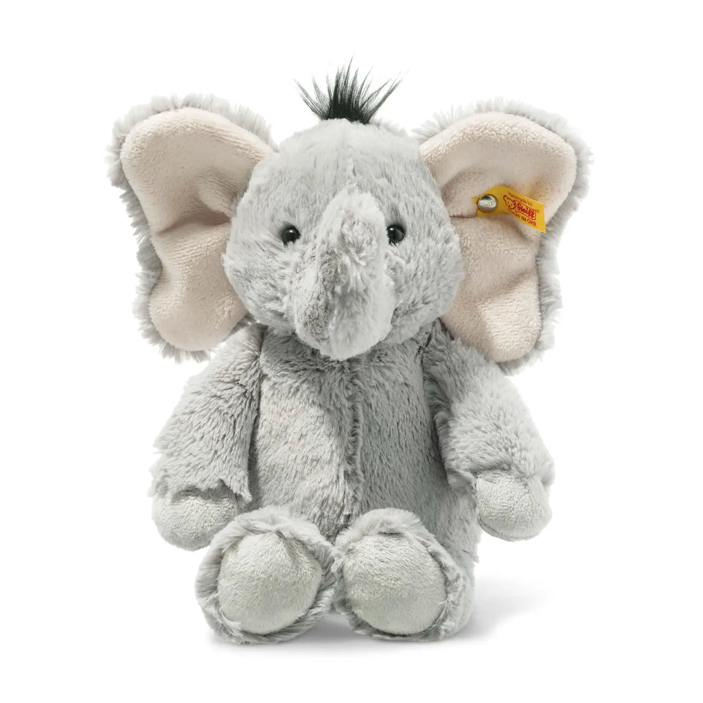 A Steiff Ella Elephant Plush Animal Toy, standing 12 inches tall, features soft gray fur, large floppy ears lined in pale pink fabric, black eyes, and a black tuft of hair on top of its head. It sits upright with a small Steiff tag attached to one ear.