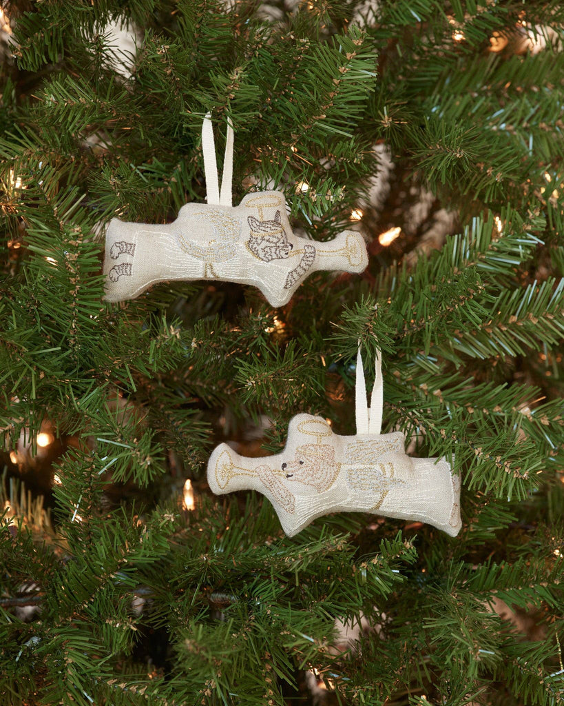 Two Angel Dog ornaments, beautifully embroidered, hang from a Christmas tree. The tree is adorned with warm white lights, creating a festive ambiance perfect for Christmas morning.