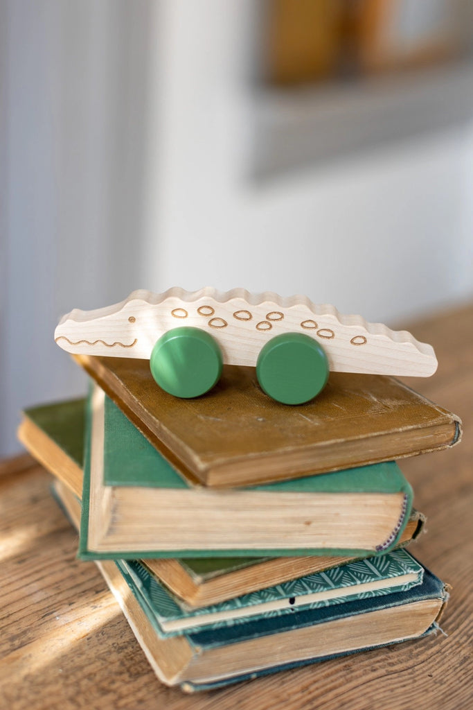 A Wooden Crocodile Push Toy with simple line details and green circular wheels sits atop a small stack of hardcover books with green and beige covers. The books are arranged haphazardly on a wooden surface. The background is blurred.