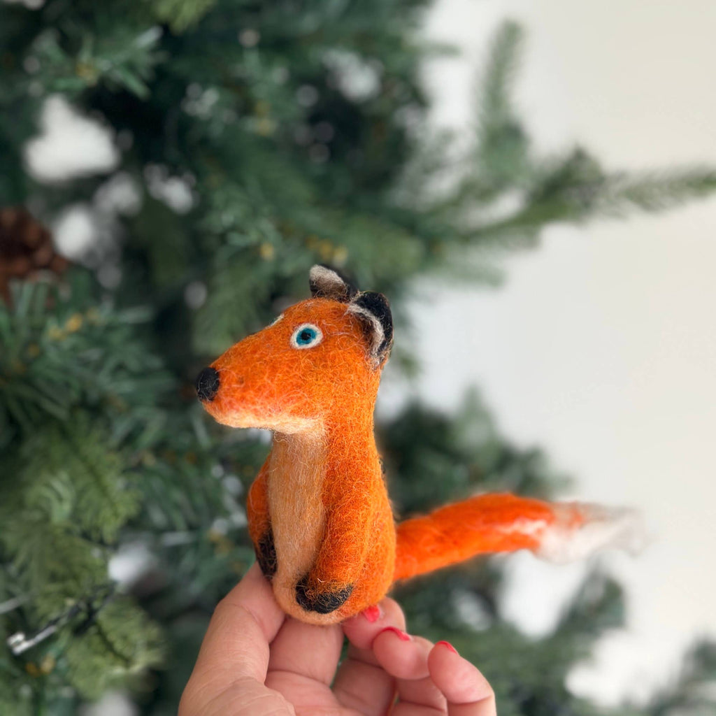 A hand holds a small, wool felt orange Fox Finger Puppet with blue eyes in front of a green Christmas tree. The fox, perfect for storytelling, features a bushy white-tipped tail and black paws.