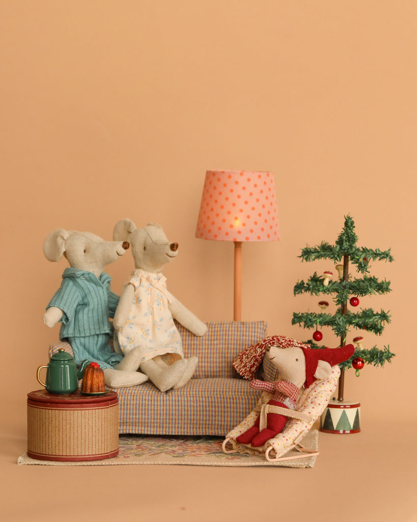 The Maileg Christmas Set features three plush mice in an inviting scene: two dressed in pastel attire seated on a plaid sofa, and another relaxing nearby with a knitted blanket. The setting includes small details such as a miniature coffee table with a teapot, a lamp, and an adorned Christmas tree. Note that it is not suitable for children under three due to choking hazards.