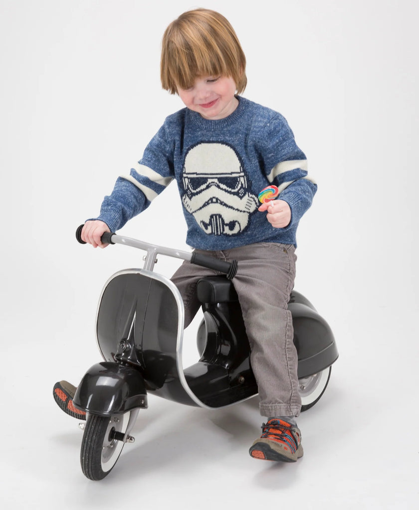 A young child with a joyful expression rides a PRIMO Ride On Kids Toy Classic, wearing a blue sweater with a classic Star Wars stormtrooper design, gray pants, and colorful shoes, on a white background.