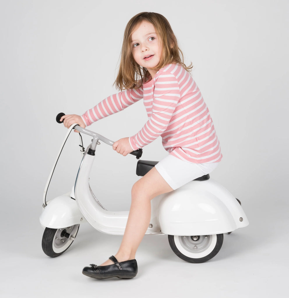 A young girl with light brown hair, wearing a pink and white striped shirt and shorts, rides a small white PRIMO Ride On Kids Toy Classic scooter, looking at the camera with a playful expression.