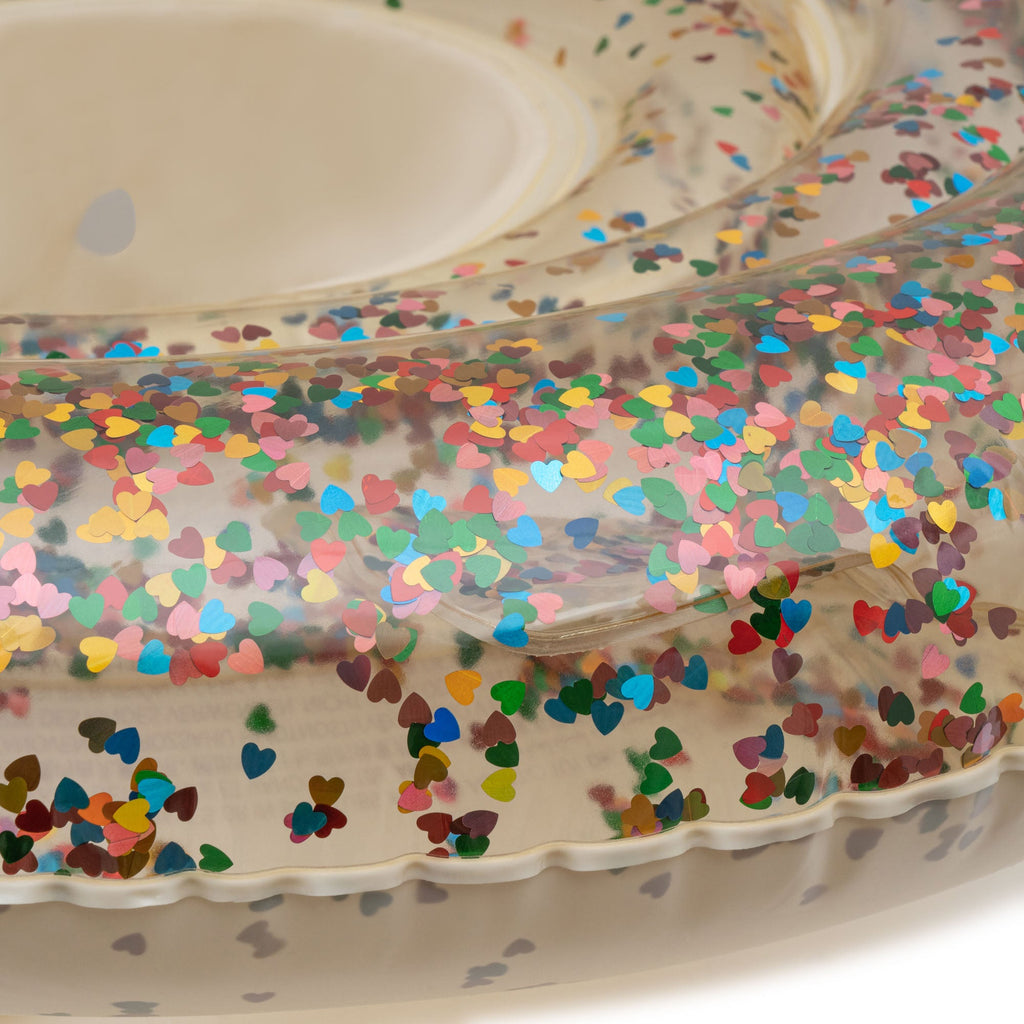 Close-up of an Inflatable Baby Swim Ring - Transparent Confetti Hearts filled with small, colorful confetti shapes. The glitter and confetti appear to be heart-shaped in various hues such as red, blue, green, yellow, and pink. The background is plain white, highlighting the float's design.