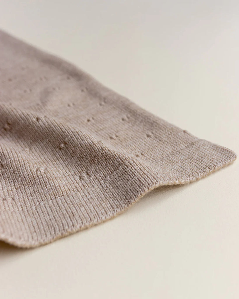 A close-up shot of a Handmade Merino Wool Bibi Blanket - Sand made from extra fine Italian merinowool, laid flat on a light surface. The texture of the fabric includes small, evenly spaced raised dots, giving it a subtle patterned appearance. The edge of the blanket is gently curved and soft.