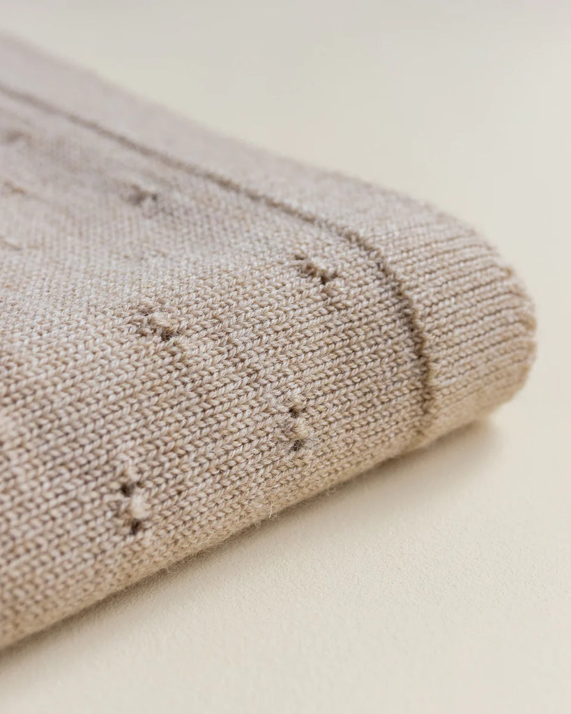 Close-up of a sand colored textured Bibi blanket folded neatly, showing detailed stitch patterns and small embroidered elements on the fabric.