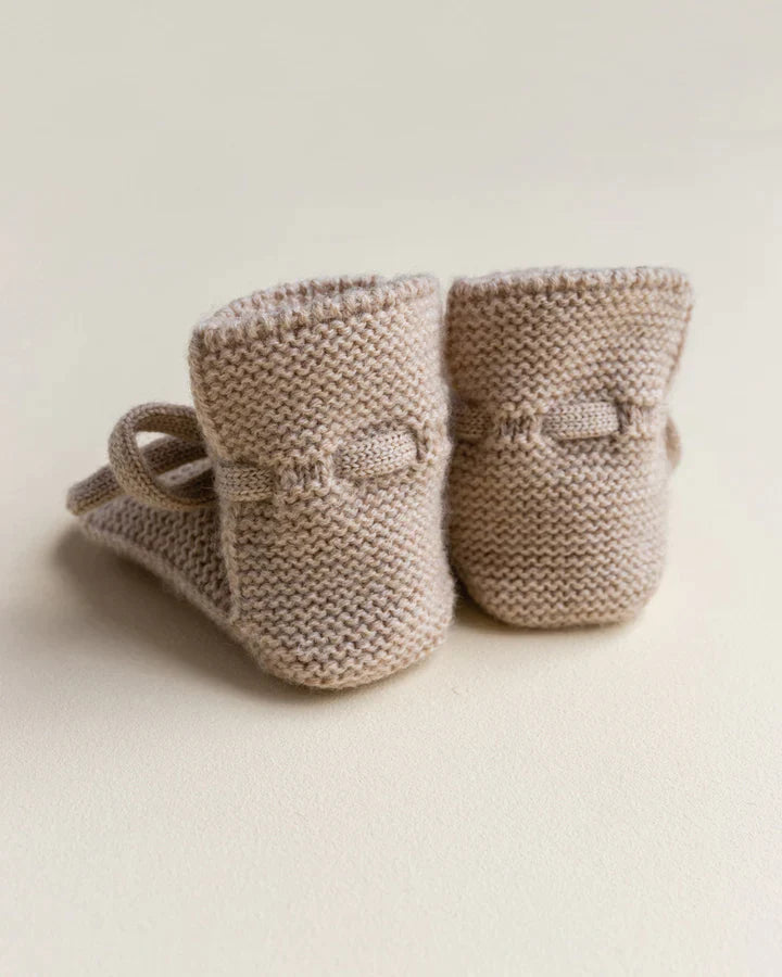 A pair of Handmade Merino Wool Booties - Sand, displayed on a light cream background. The booties feature a detailed pattern with small straps.