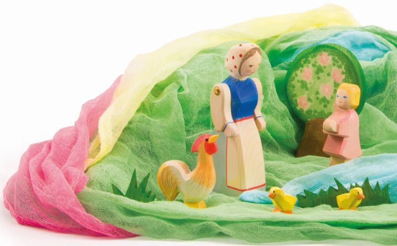 A colorful scene with handcrafted wooden toys: an adult wearing a blue dress and headscarf, a child with yellow hair, an Ostheimer Chick, Yellow, and two chicks. They stand on green terrain surrounded by pink, yellow, and blue cloth with a green tree in the background. Perfect for imaginative play.