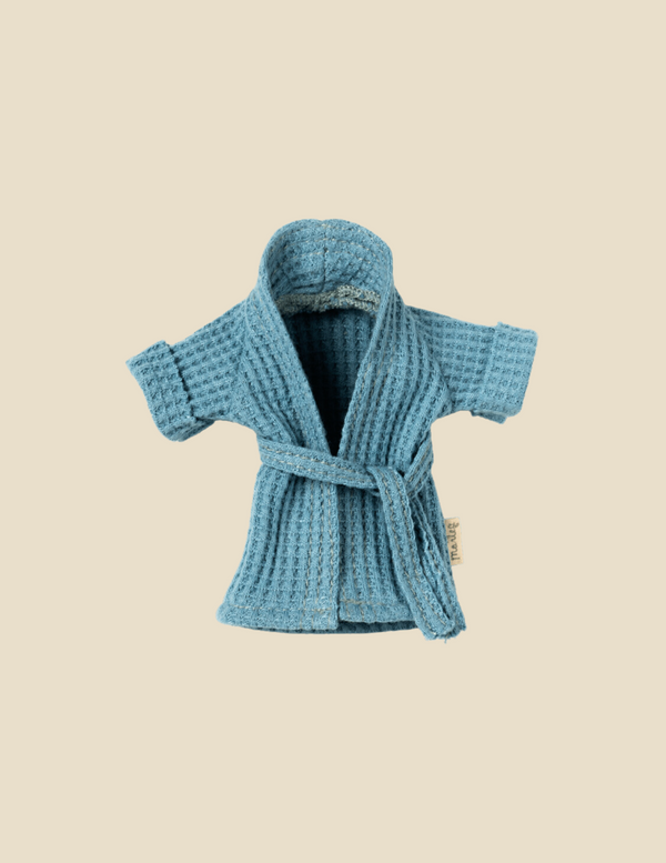 A small blue waffle-knit Maileg Extra Clothing: Bathrobe - Dusty Blue with a belt tied around the waist. The robe has short sleeves and an open front, revealing its soft, textured fabric inside. The background is a plain light beige color.