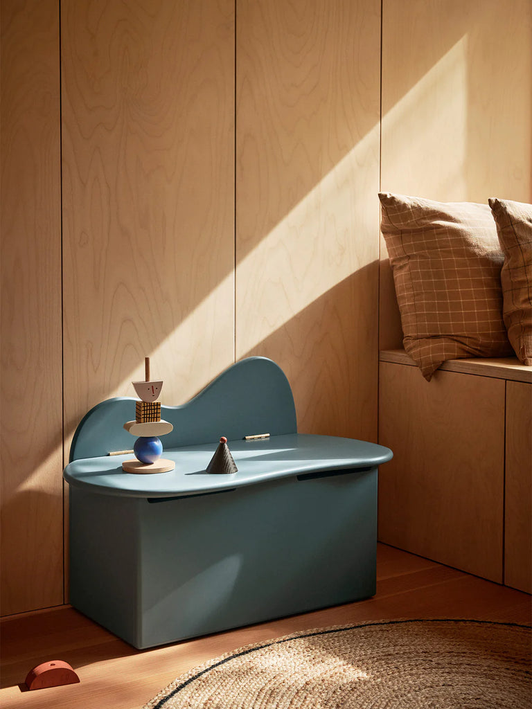 A modern, minimalist Ferm Living Slope Storage Bench - Storm with curved edges, situated in a warm, sunlit wooden room. The bench is crafted from FSC™ certified MDF and is decorated with small artistic objects.