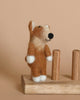 A small, needle-felted Corgi Dog Finger Puppet, featuring a standing brown and white dog with a black nose and small black eyes, is positioned on a wooden platform with two vertical wooden pegs. Handmade in Nepal, the smiling finger puppet dog contrasts beautifully against the light brown background.