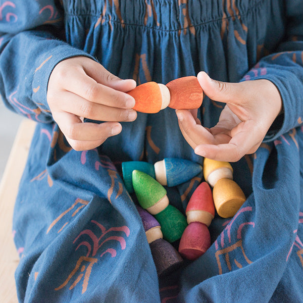 A child in a blue dress holds a vibrant, orange wooden toy piece from a Grapat Rainbow Tomten set, connecting it to another in her hands, surrounded by various colorful wooden toys.