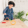 A young girl with curly hair points to the Grapat Nesting Rings on a wooden table, next to a potted plant, against a white brick wall background. She wears a blue