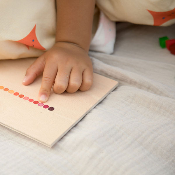 A toddler's hand touching a Grapat Mix & Match with color-matching dots, surrounded by soft, patterned cushions and scattered educational toys.