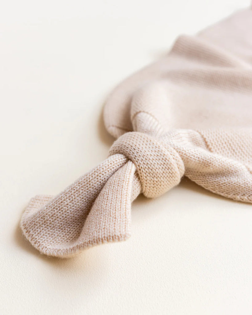 Close-up of a light beige knitted fabric tied in a neat knot. Made from luxurious Merino wool, the HVID Merino Wool Cocoon - Oat has a textured, ribbed pattern, and the knot creates a soft, casual look reminiscent of a baby cocoon. The background is a smooth, matching beige surface.