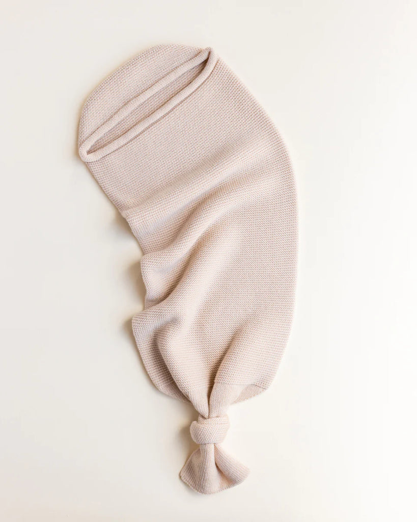 A HVID Merino Wool Cocoon - Oat laid flat on a white surface, with the lower end gathered and tied into a simple knot. Crafted from soft and textured Merino wool, it showcases a cozy and warm design reminiscent of a baby cocoon.