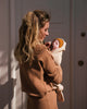 A woman with long blond hair stands holding a sleeping baby cocooned in an HVID Merino Wool Cocoon - Oat and wearing a yellow knit hat. They are in front of a closed door, and the woman is dressed in a matching warm-colored outfit. Sunlight casts shadows behind them, illuminating their serene moment.