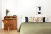 A minimalist bedroom featuring a neatly made bed with a green blanket and a embroidered Coral & Tusk Great Grey Owl Pillow, a wooden writing desk with books, and a chair, framed abstract art on the wall, and simple white walls.