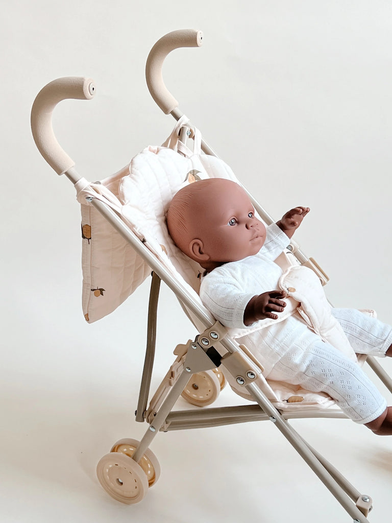 A baby doll with medium skin tone is strapped into a Konges Sloejd Doll Stroller - Lemon with a padded seat and hood. The stroller features dual handles and double wheels for smooth movement. The doll, dressed in a white outfit and securely fastened by a harness, faces forward against a plain white background.