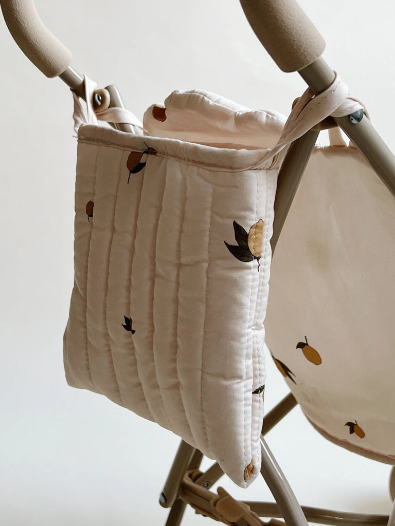 A close-up of a padded, quilted bag hanging from a beige Konges Sloejd Doll Stroller - Lemon frame. The bag is cream-colored with a pattern of small, illustrated lemons and leaves. The background is plain and white, highlighting the stroller's double wheels for added stability.