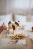 A close-up of wooden farm animal figurines, handcrafted linden wood toys, on a light-colored fabric surface. A Handmade Wooden Cow, black-and-white, and another brown cow made with child-safe paint stand near a small pile of hay, with a blurred white rooster and other indistinct figures in the background.