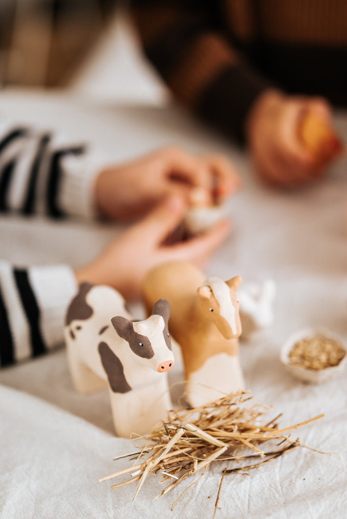 Two Handmade Wooden Cows, one brown and one black and white, stand on a white surface with a small pile of straw in front of them. In the background, two blurred figures, one in a striped shirt, interact closely while holding small objects. These toys are handmade in Serbia using child-safe paint.