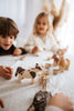 Two children, one with brown hair and the other with blonde hair, play with handcrafted linden wood toys on a white tablecloth. The Handmade Wooden Cow is painted with child-safe paint. Hay and small bowls are placed around it.