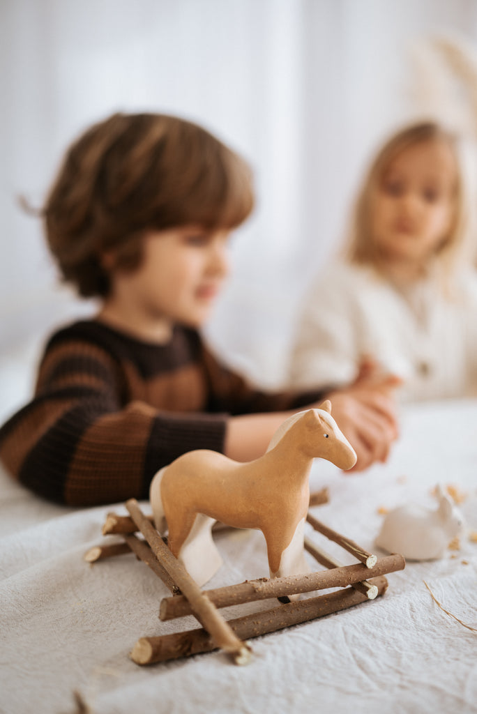 Two children are sitting at a table focusing on an activity. In the foreground, there's a Handmade Wooden Horse on a wooden frame made of linden wood, painted with child-safe paint. The child with brown hair is wearing a striped sweater, and the child with blonde hair is slightly blurred in the background.