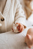 A child in a cream-colored, knitted sweater holds a Handmade Wooden Rabbit on a cloth-covered table. Only the child's hand and part of their torso are visible. The scene appears warm and cozy. The figurine is hand-painted with non-toxic paint.