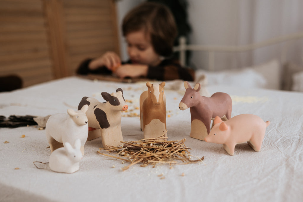 A young child in the background is focused on an activity at a table, while several handcrafted wooden animal figurines, including a Handmade Wooden Horse, donkey, pig, and rabbit painted with child-safe paint, are arranged on a cloth surface in the foreground, surrounding a small pile of hay.