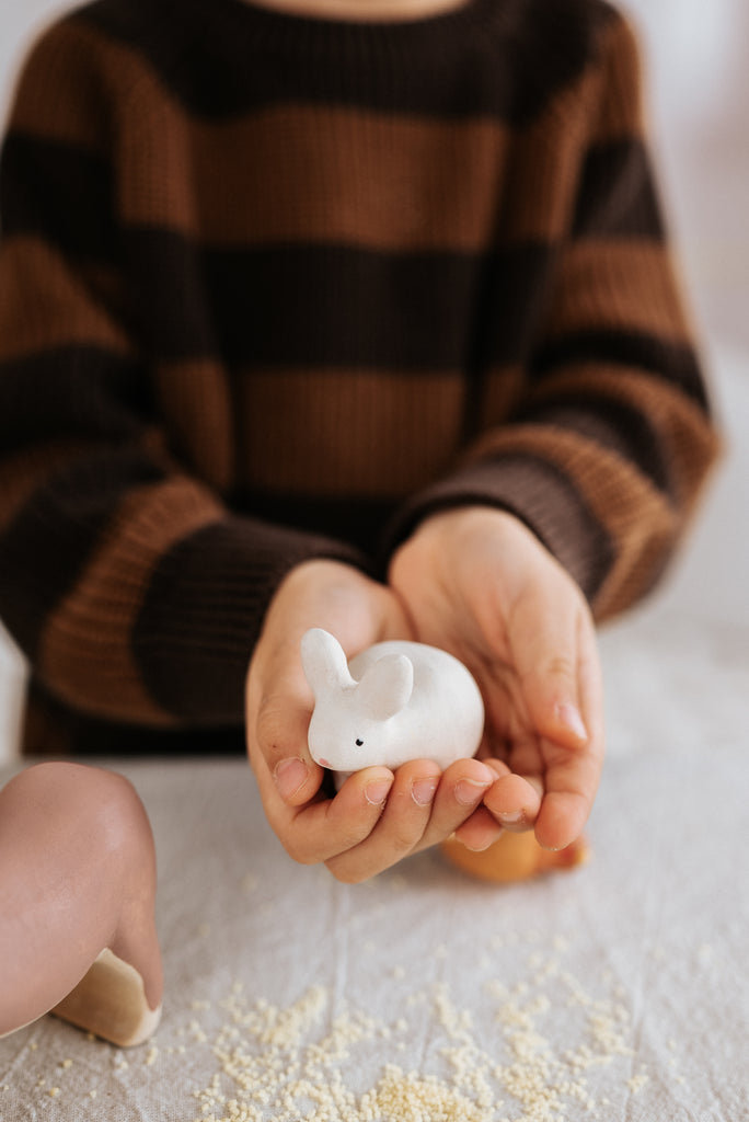 A person in a brown and black striped sweater gently holds a Handmade Wooden Rabbit in their hands. Handcrafted wood toys handmade in Serbia, the focus is on the hands and figurine, with a soft background. Some scattered crumbs are visible on a white surface below.