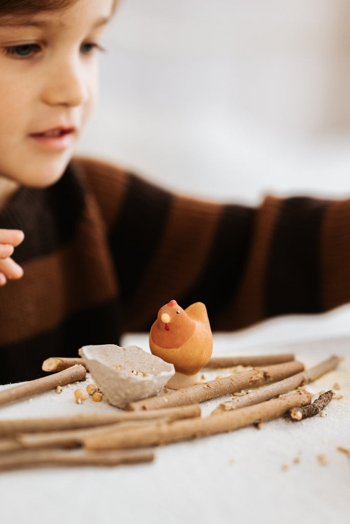 A child in a brown and black striped sweater intently examines the Handmade Wooden Chicken, a small figurine crafted from linden wood. Resting on a fabric surface with scattered twigs, this handcrafted toy features child-safe paint and captures the child's complete focus.