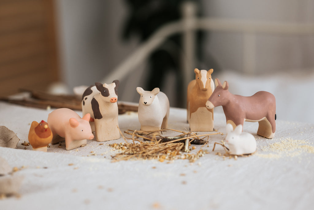 The Handmade Wooden Chicken, crafted from linden wood and coated with child-safe paint, is displayed on a white surface. Accompanying the chicken are other farm animal figurines such as a cow, pig, sheep, horse, donkey, and rabbit. The scene is completed with hay and scattered grains in the foreground to evoke a rustic farm atmosphere.