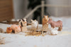 A Handmade Wooden Cow is arranged on a white cloth with other small, handcrafted linden wood farm animal toys. The set includes a pig, sheep, goat, donkey, chicken, and rabbit. Some straw and scattered seeds are on the cloth around the animals. The background is softly blurred.