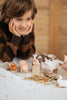 A young child with brown hair and wearing a striped sweater rests their chin on their hands while looking intently at a Handmade Wooden Cow, part of a set that includes other handcrafted linden wood farm animal figurines like a pig, chicken, and sheep—each painted with child-safe paint and arranged on a table scattered with hay.