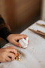 A child holding a small Handmade Wooden Rabbit, handcrafted wood toys handmade in Serbia, while resting their hand on a light-colored tablecloth. Scattered pieces of nuts and a few sticks are placed on the table. The child is wearing a dark brown and beige sweater.