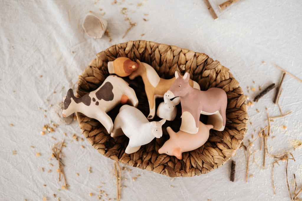 A woven basket holds a Handmade Wooden Cow and four other small wooden farm animal figurines, including a horse, donkey, sheep, and pig. These handcrafted linden wood toys are painted in natural, child-safe colors and are placed on a white cloth with scattered straw and wooden pieces around. Handmade in Serbia.