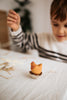 A child joyfully plays with a Handmade Wooden Chicken, artfully perched on an eggshell, while wearing a striped sweater. The softly blurred background features pieces of straw scattered across the table, enhancing the charm of its linden wood craftsmanship.