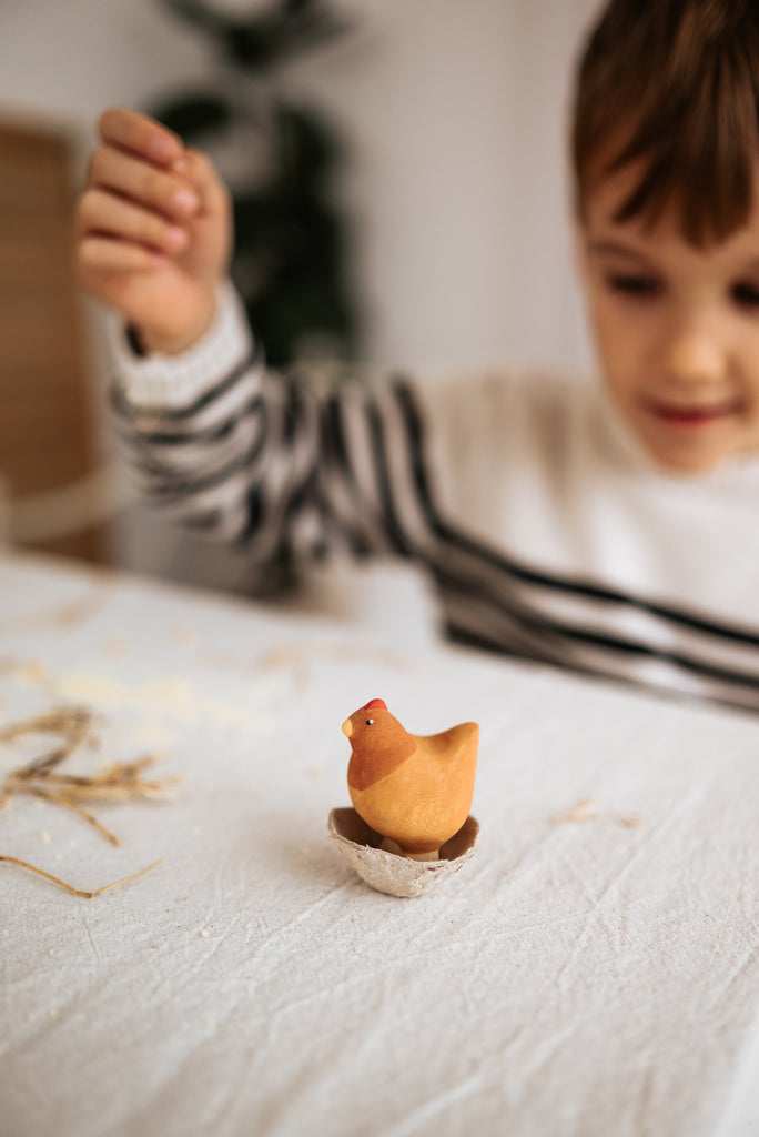 A child joyfully plays with a Handmade Wooden Chicken, artfully perched on an eggshell, while wearing a striped sweater. The softly blurred background features pieces of straw scattered across the table, enhancing the charm of its linden wood craftsmanship.