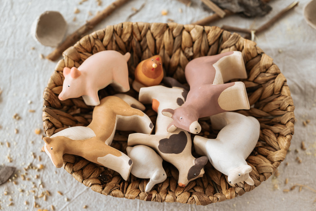 A textured surface features a woven basket filled with ceramic animal figures, including cows, horses, and the Handmade Wooden Chicken. Scattered grains and linden wood sticks adorn the scene, while child-safe paint ensures these handcrafted toys are both safe and charming.