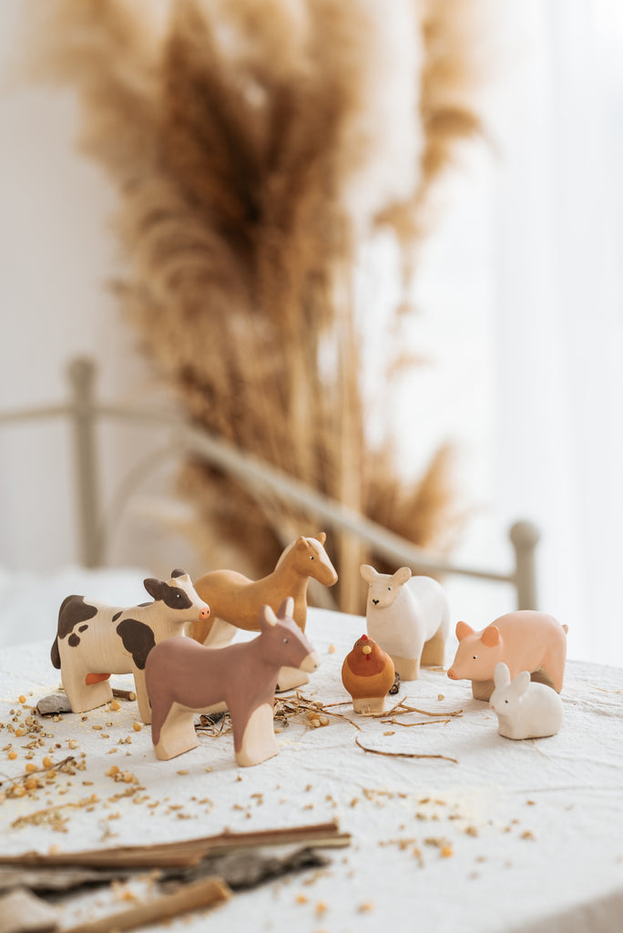 A Handmade Wooden Horse, handcrafted from linden wood and painted with child-safe paint, is placed on a white tablecloth. Wheat stalks blur in the background while scattered grains and sticks add a rustic feel to the scene.