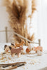 A Handmade Wooden Rabbit, including a cow, horse, sheep, pig, hen, and duck, are placed on a table covered with straw and grain. Handmade in Serbia using non-toxic paint, these charming farm animals sit against a backdrop of dried pampas grass decor and blurred furniture for a cozy indoor setting.