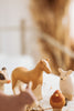 A Handmade Wooden Horse, along with small, handcrafted linden wood farm animal figurines including a cow, sheep, and chicken painted with child-safe paint, is arranged on a light wooden surface. The background is blurred, creating a soft and warm ambiance.