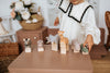 A child in a white dress with black trim plays with a Handmade Wooden Rhinoceros on a brown table. The figures, painted with non-toxic paint, include a zebra, rhino, elephant, giraffe, hippopotamus, and crocodile. Handmade by Meshka and Friends, they sit against floral decor and wicker baskets.