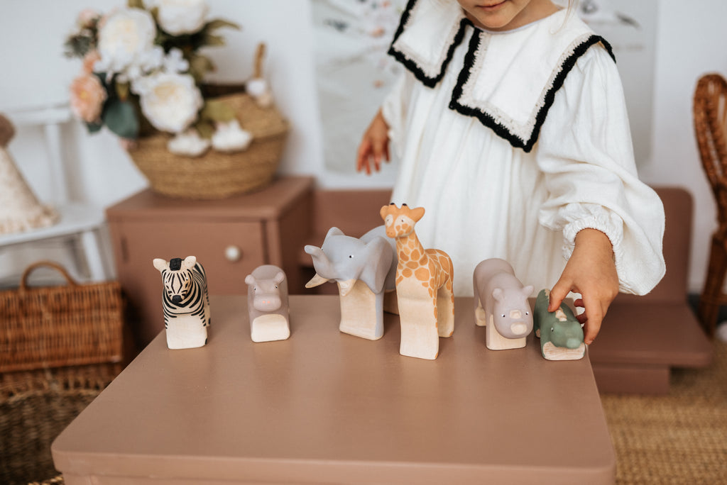 A child in a white dress with black trim plays with a Handmade Wooden Rhinoceros on a brown table. The figures, painted with non-toxic paint, include a zebra, rhino, elephant, giraffe, hippopotamus, and crocodile. Handmade by Meshka and Friends, they sit against floral decor and wicker baskets.