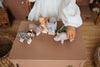 A child wearing a white long-sleeved shirt is playing with a handmade wooden rhinoceros on a small, tan table. The figurine, painted with non-toxic paint, is accompanied by others including a zebra, giraffe, hippos, and a crocodile. Wicker baskets and a brown cabinet are in the background.