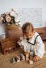 A young child with light brown hair is playing on a woven rug with handcrafted linden wood toys, including an elephant, giraffe, and Handmade Wooden Rhinoceros. Behind them, there is a wicker basket, a basket of flowers, and artwork with a floral design on the wall. The toys are handmade by Meshka and Friends.