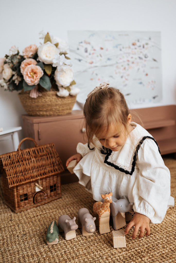 A young child with light brown hair is playing on a woven rug with handcrafted linden wood toys, including an elephant, giraffe, and Handmade Wooden Rhinoceros. Behind them, there is a wicker basket, a basket of flowers, and artwork with a floral design on the wall. The toys are handmade by Meshka and Friends.